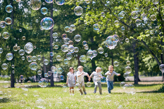Green public park illuminated with sunbeams, colorful soap bubbles flying in air, group of little children playing active games