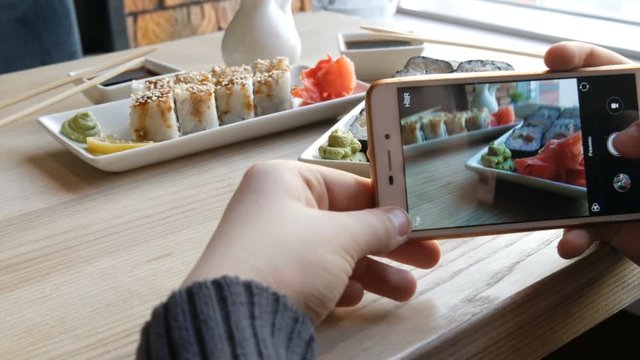 Hands of a teenager make a photo of food on a smartphone. A set of sushi rolls from Japanese cuisine on the background of porcelain teapot for soy sauce and saucer in a stylish cafe