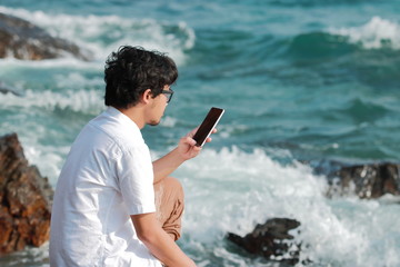 Young Asian man with mobile smart phone sitting on the rock of natural seashore. Summer lifestyle concept.