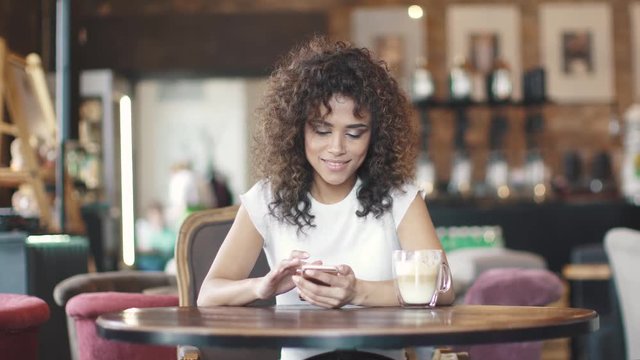 Portrait of a beautiful hispanic girl in a cafe. girl texting message on smartphone and smiles