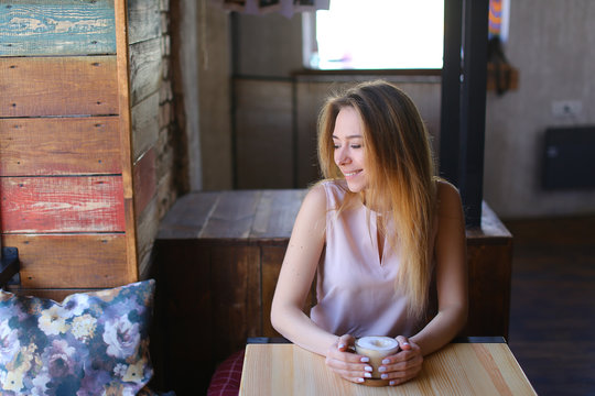Young woman decided to devote time to herself on weekend and went to coffee house, smiling fair-haired female wearing white blouse sitting near wooden table with glass cup of cappuccino on cork saucer