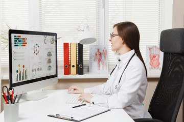 Young woman sitting at desk, working on modern computer with medical documents in light office in...