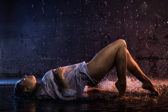Girl in the white shirt with water drops and dark walls background illuminated by light during a photoshoot with water