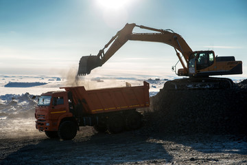 Against the background of a snowy arctic landscape, the excavator loads crushed stone into the body of an orange dumper..