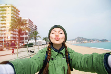 Obraz premium Pretty young female tourist takes travel selfie at the beach while spring vacation Barcelona in Spain.Beautiful young brunette making selfie sending cute air kiss.Beautiful young travel blogger.