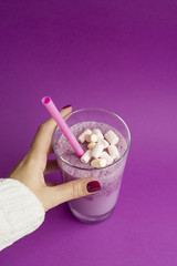 Female hands holds glass of berry smoothie with straw on purple background. Pink milkshake with copy space. Healthy drink. Blueberry cocktail and marshmallow.
