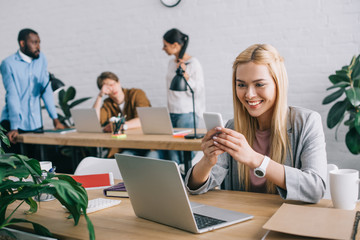 happy businesswoman typing on smartphone and colleagues standing behind in modern office