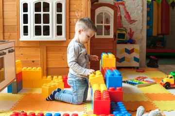the boy is 4 years old, the blond plays on the playground indoors, builds a fortress from plastic blocks