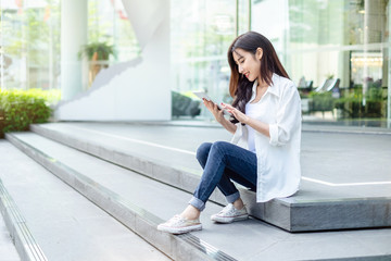 Young and beautiful Asian woman using her tablet PC while sitting on the steps outside the modern office building in the urban city