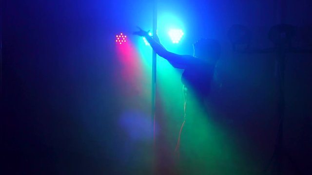 Young woman's silhouette dancing on pole in smoke. Close-up silhouette of a sexy girl near a pole on a background of bright colored spotlights in a nightclub. Pole dance.