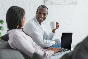 Fototapeta na wymiar african american girlfriend using laptop and boyfriend drinking coffee at home