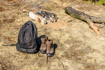 Backpack and touristic boots on a ground in a coniferous forest. Bonfire on a background