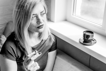 young woman smokes at the window. concept of loneliness and depression. portrait in flat, apartment - obrazy, fototapety, plakaty