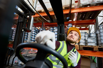Low angle portrait of young warehouse worker sitting inside forklift moving goods from tall storage...