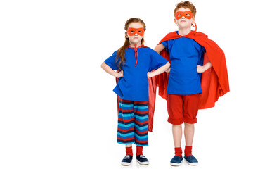 super children in masks and cloaks standing with hands on waist and looking at camera isolated on white