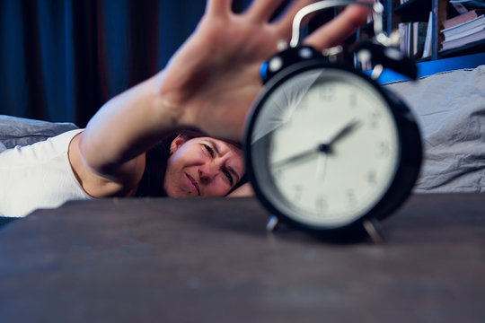 Portrait of dissatisfied woman with insomnia stretching arm to alarm clock at night