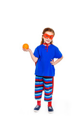 adorable child in superhero costume holding orange and smiling at camera isolated on white