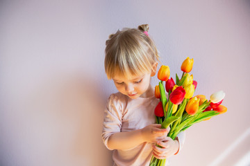 Obraz na płótnie Canvas A serious little blond-haired girl is holding a bouquet of colorful tulips in hands in a sunny room at home. Holy concept.