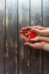 Two pairs of hands, men and children on a wooden background. Four hands are holding a red, bright heart.