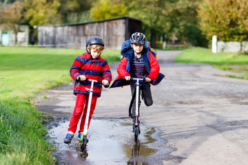 Fototapeta na wymiar Two little kids boys riding on push scooters on the way to or from school. Schoolboys of 7 years driving through rain puddle