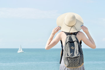 Fototapeta na wymiar Girl in a hat with a backpack standing on the coastline. Sailboat in the distance. Back view
