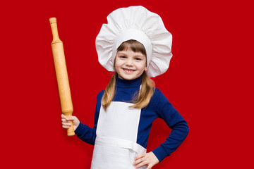 Happy little girl in chef uniform holds rolling pin isolated on red. Kid chef. Cooking Process Concept