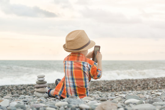 Little boy takes pictures on the smart phone. Sits on a pebble beach