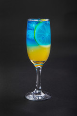 A multi-colored, two-layered yellow and blue cocktail in a high glass with ice cubes with the taste of pineapple, orange and a slice of lemon. Side view. Isolated black background. Drink for the menu