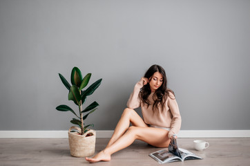 beautiful young brunette girl in white short shorts sitting on floor next to ficus on gray background in drinks coffee