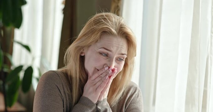 Woman smiling to herself in her anxiety and then running her hands through her long blond hair as she rocks to and fro in a chair.