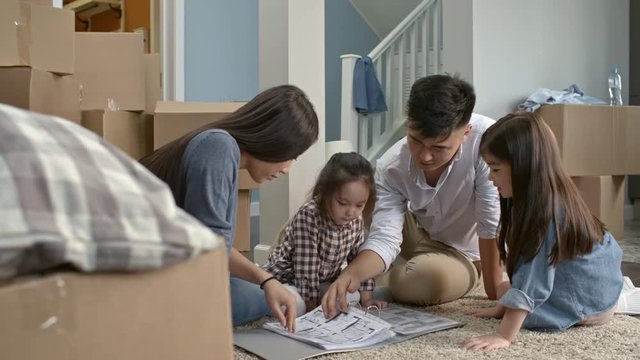 PAN of happy Asian family sitting on floor and looking through folder with layout plan of their new house; unpacked cardboard boxes lying on floor