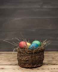Easter eggs of different colors in a wicker basket with hay on a wooden background