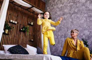The mother sitting on the bed and daughter jumping on the bed