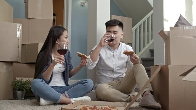Zoom out shot of happy Asian couple sitting on floor of their new apartment and relaxing after moving house: they eating pizza and drinking wine