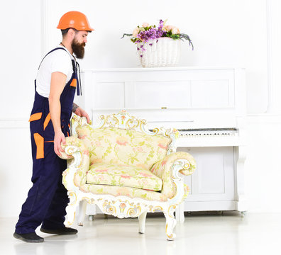 Courier delivers furniture in case of move out, relocation. Man with beard, worker in overalls and helmet lifts up armchair, white background. Delivery service concept. Loader carries armchair.