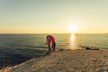 Young woman doing yoga on a rocky seashore at sunset. The concept of a healthy lifestyle. Harmony. Human and nature. The background of the blue ocean.