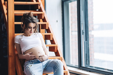 Beautiful saucy pregnant woman with long brown hair in the white t-shirt and blue jeans, portrait of beautiful pregnant woman, cute pregnant belly, future mothers