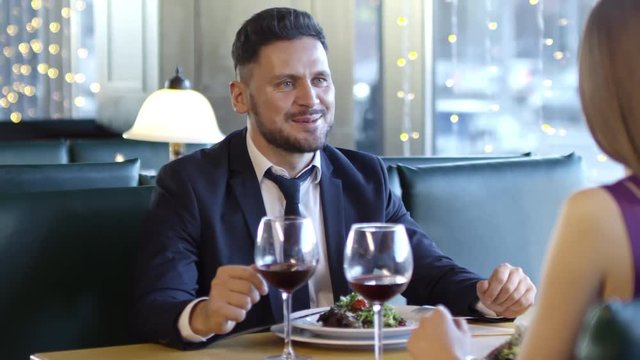 PAN of bearded man in suit and his unrecognizable female date sitting at table and drinking while having dinner in restaurant