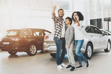 African american family at car dealership. Mother, father and son are taking selfie in front of new...