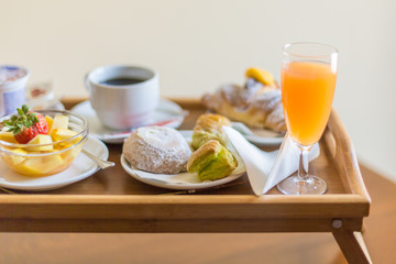 Breakfast on a wooden tray. Green puff pastry, bowl with fruit, bun with icing sugar, coffee cup, croissant, glass of orange juice