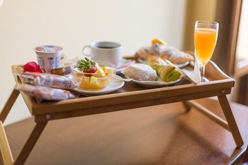 Breakfast on a wooden tray, detail of a glass bowl with fruit. Glass of orange juice, buns, croissant, puff pastry, coffee cup and yogurt