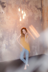 Photo of cheerful redhead young woman in mustard dress and jeans standing over grey wall background. Blur effect.