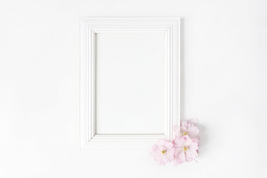 White blank wooden picture frame mockup with pink Japanese cherry blossoms lying on the white table. Poster product design. Styled stock feminine photography. Home decor. Spring concept.