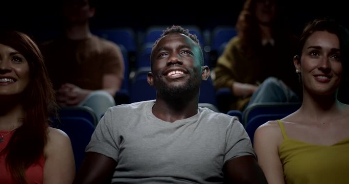 A young man siting in a movie theatre smiling, enjoying a movie