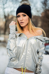 Portrait of attractive blonde woman wearing hat and silver jacket looking at camera while posing on blurred winter park background