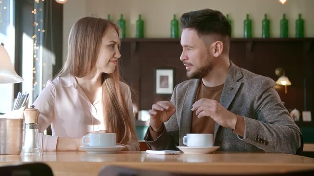 Medium shot of cheerful young woman and man with beard sitting at table in cafe and chatting over coffee