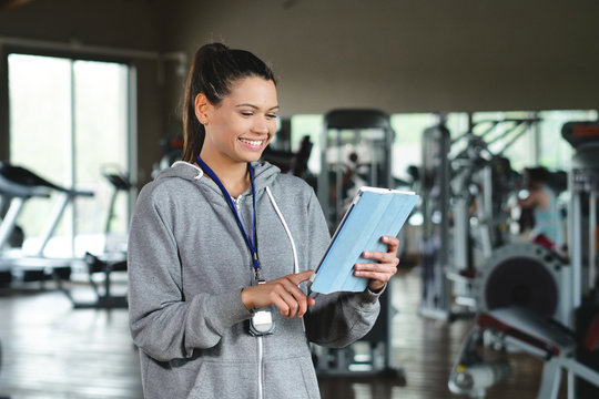 A young personal trainer (girl) holds an Ipad with the gym in the background. Concept: Sport, fitness, happiness.