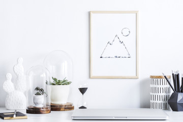 The creative desk with mock up poster frame, laptop, cacti, hourglass nad office accessories. Space of freelancer.