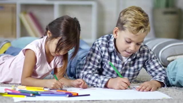 Tracking shot of two cute schoolchildren lying on the floor and drawing pictures with colored felt-tip pens and pencils