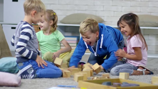 Group of four playful primary school children sitting in circle around toy building and destroying it, toy blocks falling down